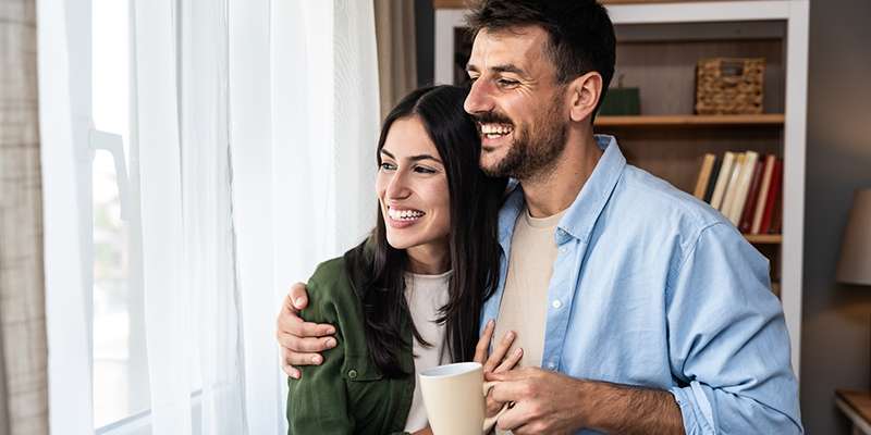 dreamstime319662356_crop Smiling young couple standing in front of a window. He is holding a coffee mug and has his arm around the woman. They are both looking out the window and smiling.